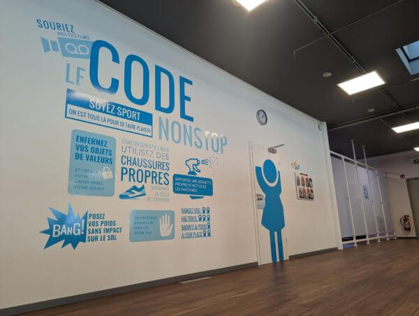 Décoration murale intérieure d’une salle de sport avec lettrage bleu Le Code Non Stop et illustrations rappelant les règles d’hygiène et de sécurité
