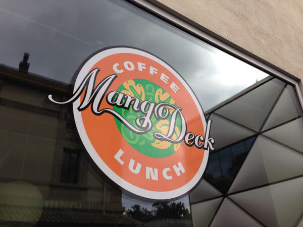 Logo circulaire Mango Deck apposé sur une vitrine avec texte Coffee Lunch et illustration de mangue stylisée au centre